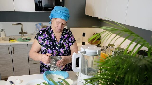 Grandmother with headscarf holding hand mixer and metal flour sifter over blue bowl. Home baking and step by step dough preparation in modern kitchen.