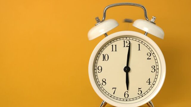 A classic white twin bell alarm clock stands against a cheerful yellow backdrop. Its hands point to six o'clock, creating a sense of early morning or evening.