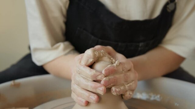 Close up shot of unrecognizable hands of woman in overalls creating cone shape with wet clay on potters wheel in workshop