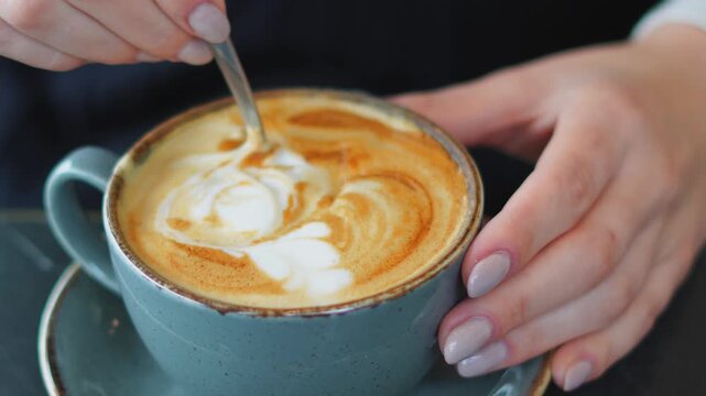 Woman stirs coffee in blue cup, creating latte art with spoon in cafe setting