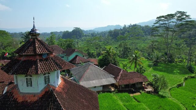 Aerial drone footage in Mekarmulya, Pasirkuda, Cianjur regency, Indonesia, with a small village and a colorful mosque, a wide green rice field valley, and mountains behind