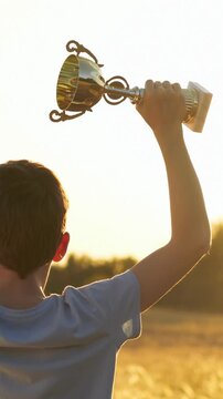 Teenager celebrates victory with trophy in sunset. Boy raises golden award, showing excitement and joy. Beautiful outdoor background enhances vertical scene. Successful teenager holds cup in his hands