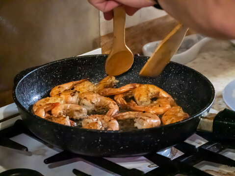 Cook frying shrimp in pan on home stove. Person using wooden spatula to stir fresh seafood. Process of cooking healthy protein meal in kitchen. Culinary hobby and gastronomy concept.