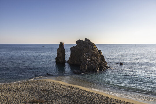View of craggy rocks rise majestically from the turquoise sea, framed by a sandy beach under a clear sky, Monterosso al Mare, Cinque Terre, Liguria, Italy.
