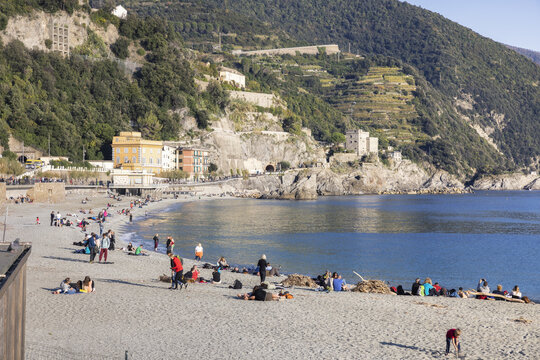 View of the beach filled with people relaxing and sunbathing under the clear sky, set against the dramatic backdrop of lush mountains, Monterosso al Mare, Cinque Terre, Liguria, Italy.