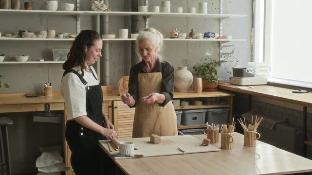 Elderly craftswoman teaching young apprentice how to cut clay with string at workbench in modern pottery studio
