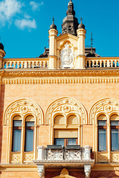 View of an ornate, sun-kissed building facade rises under a vibrant blue sky, showcasing intricate architectural details, Zmaj Jovina, Novi Sad, Vojvodina, Serbia.