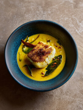 A piece of crispy-skinned white fish served in a bright yellow turmeric and coconut milk sauce with curry leaves in a blue ceramic bowl.