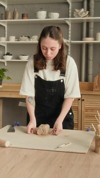 Vertical shot of young woman wearing dark jumpsuit, cutting portions of light brown clay with string, leaning over work surface in studio
