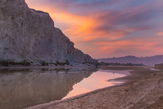View of the river mirroring the vibrant twilight sky, reflecting the rugged cliffs and sandy banks, a serene landscape, Hingol National Park, Balochistan, Pakistan.