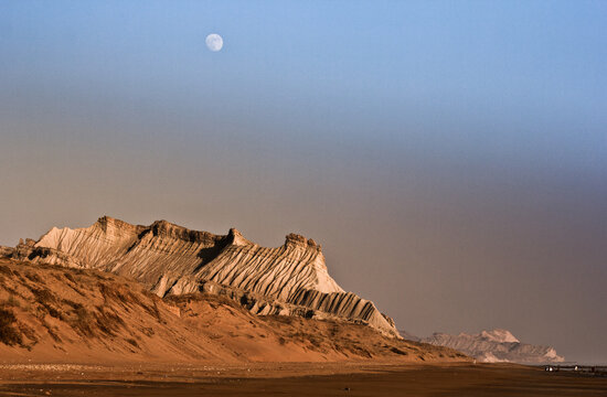 View of the arid landscape under a pale moon, where wind-sculpted cliffs meet sandy shores, creating a stark contrast of textures and tones, Hingol National Park, Balochistan, Pakistan.