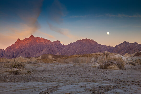 View of jagged peaks kissed by fiery sunset hues, casting long shadows across the arid landscape under the watchful gaze of a full moon, Hingol National Park, Balochistan, Pakistan.