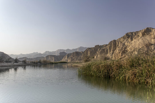 View of tranquil waters reflecting the rugged cliffs and sparse vegetation under a pale sky in Hingol National Park, Hingol National Park, Balochistan, Pakistan.