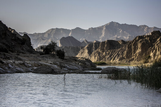 View of rugged cliffs and sparse vegetation meet the tranquil waters of a river, framed by distant mountains in Hingol National Park, Hingol National Park, Balochistan, Pakistan.