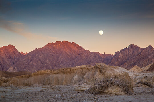 View of rugged, sunlit mountains under a twilight sky, with a full moon casting a soft glow over the arid landscape, Hingol National Park, Balochistan, Pakistan.