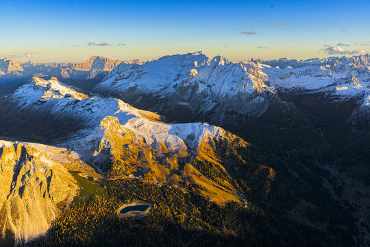 Aerial view of jagged, snow-dusted peaks kissed by golden sunlight, contrasting with the deep shadows in the valleys below, Marmolada, Trentino-South Tyrol and Veneto, Italy.
