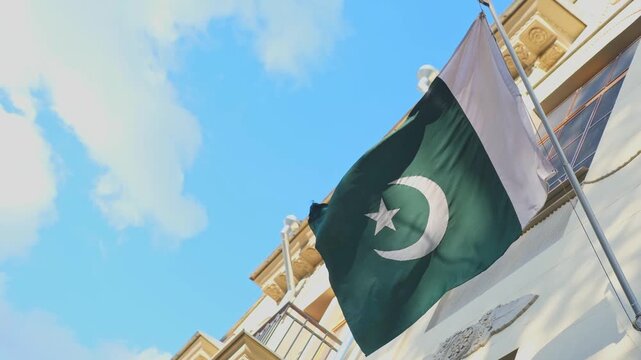Pakistani flag swaying and fluttering in wind against blue sky with clouds and historical buildings. Flag of Pakistan. Flag day celebration, patriotism concept. Embassies and consulates of countries