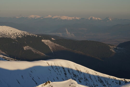 Aerial view of snow-capped peaks and forested hills create a stark contrast against the distant, hazy horizon, Nizke Tatry, Zilina Region, Slovakia.