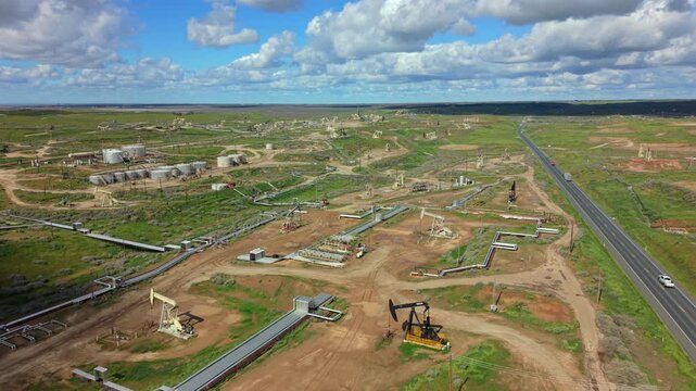 Wide aerial view of an active oil field with pumpjacks pipelines and service roads, industrial energy production infrastructure across a vast rural landscape under dramatic cloudy sky. California