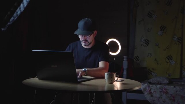 A man in black clothes sits at a table. He wears a baseball cap and glasses. He works remotely. Freelancer. Night. The lights are on. There's a laptop on the table. A ceramic mug. Home interior. Stora