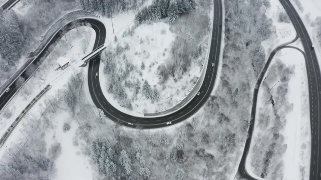 Cinematic aerial drone shot of a winding mountain road with cars driving through a snowy forest while a train crosses the scene on a bridge. Multi-layer transportation movement in a winter landscape