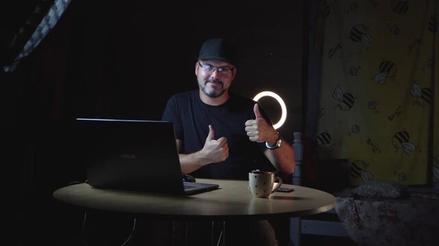 A man in black clothes sits at a table. He wears a baseball cap and glasses. He works remotely. Freelancer. Night. The lights are on. There's a laptop on the table. A ceramic mug. Home interior. Stora