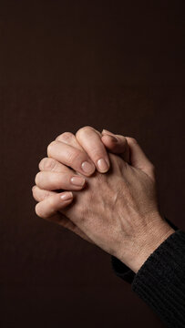 Close-up of clasped, mature Caucasian person hands, in prayer or deep contemplation against a dark, somber background. This image evokes themes of faith, hope, solace, and spiritual reflection