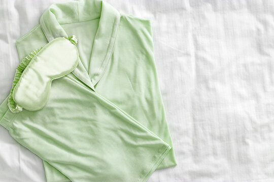 Top view of folded pastel green woman pajama with satin sleep eye mask on white bed. Minimal flat lay cozy comfort pyjamas for sleeping and relaxed. Aesthetic lifestyle photo of stylish sleepwear