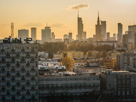 Aerial view of a sprawling cityscape punctuated by modern skyscrapers piercing through the golden haze, contrasting against the older buildings below, Warsaw, Poland.