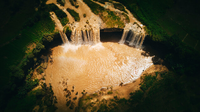 Aerial view of muddy water cascading down the rocky cliff into a circular pool, framed by vibrant green vegetation, Batadon, Kaduna, Nigeria.