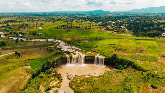 Aerial view of cascading waterfalls plunge into a pool amidst lush green fields, reflecting the sky above, Batadon, Kaduna, Nigeria.