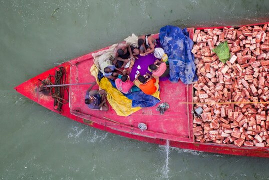 Aerial view of a red boat carrying a pile of bricks and people, creating a striking contrast against the murky water, Munshiganj, Dhaka Division, Bangladesh.