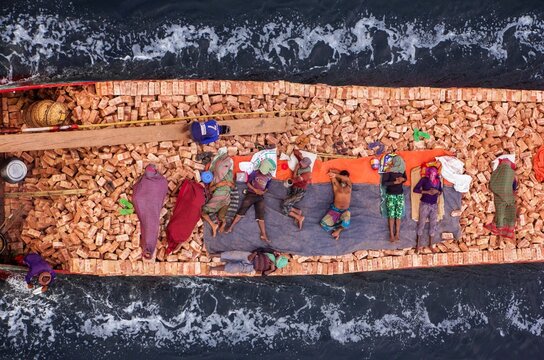 Aerial view of laborers resting on a barge laden with bricks, contrasting the rough texture of the cargo with the serene flow of the river, Dhaka, Dhaka Division, Bangladesh.