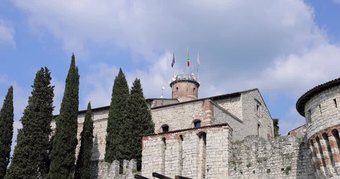 Brescia castle arch and drawbridge beams with cypress and flag tower, low angle camera tilts up along rugged stone walls to Torre Mirabella where flags flutter in summer breeze beneath drifting clouds
