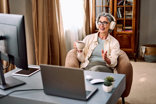A beautiful middle-aged woman wearing headphones and glasses sits at a desk in front of a monitor and a laptop, attending an online business meeting at home. Concept of remote work.