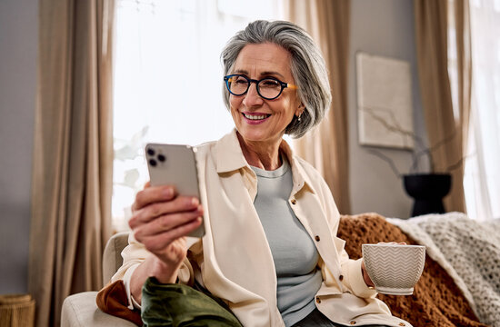 Portrait of a beautiful smiling middle-aged gray-haired woman wearing glasses, using a phone while sitting in a cozy living room with a cup of coffee or tea.