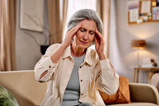 Elderly woman sitting on couch with eyes closed, holding temples, appearing to have a headache or discomfort.