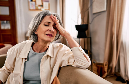 Middle-aged woman with gray hair sitting on sofa at home holding head in pain, stressed and fatigued expression, healthcare and mental health concept.