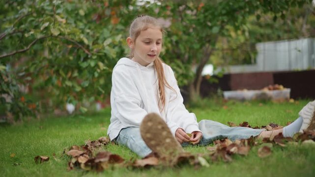 Young girl sitting on grass collecting leaves, wearing white hoodie and jeans, braided hair, soft smile, suburban garden backdrop, crisp fall light, candid backyard daydreamer vibe