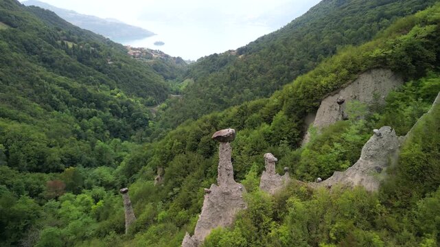 Drone flyover of Zone earth pyramids with boulder caps, drifting toward Lake Iseo over green woodland in Lombardy Italy highlighting alpine erosion landforms