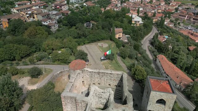 Drone pullback over Padenghe sul Garda Castle, camera drifts backward from crenellations to a wide panorama of tiled roofs olive groves and curving highway through trees