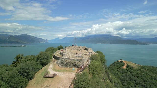 Drone rises above Rocca di Manerba del Garda on a sunny summer day, sweeping over cliffs and ancient ruins to reveal Lake Garda bays and distant mountains