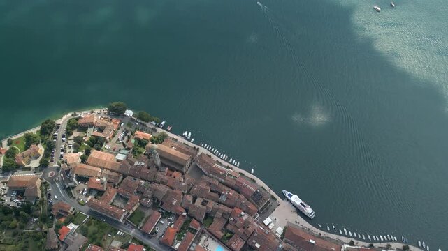 Drone reverse tracking shot over Salo waterfront on Lake Garda, flying backward with camera tilted down to reveal terracotta roofs church dome promenade and ferry dock beside rippled blue water