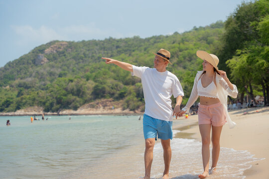 Happy couple walking on tropical beach holding hands, man pointing forward. Summer vacation travel lifestyle, love and relationship, seaside landscape, freedom and exploration concept