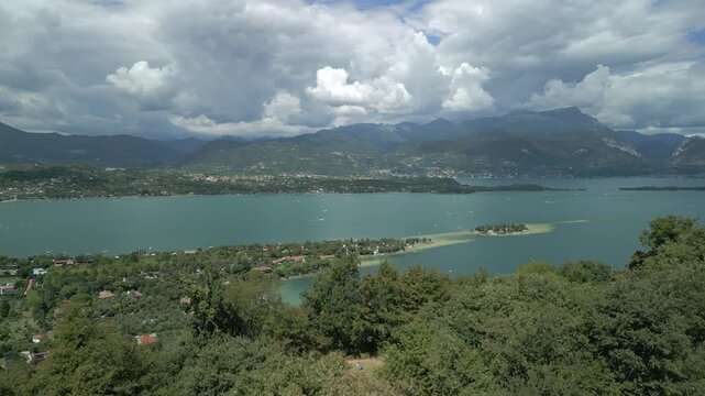 Hovering drone pans left to right above Rocca di Manerba ruins, revealing Lake Garda mountains and San Biagio Island (Isola dei Conigli) in sunny summer light