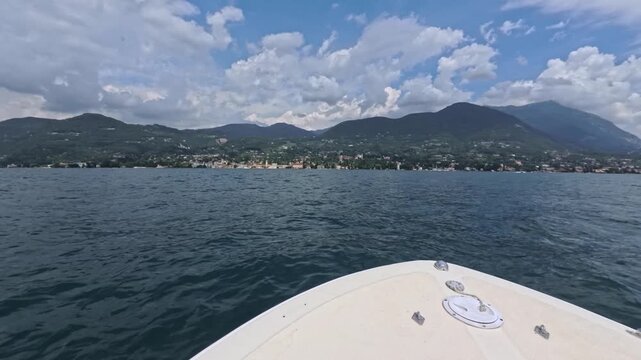 Motorboat bow gliding across Lake Garda toward alpine foothills, view shows choppy blue surface and a hillside village framed by forests and high peaks in summer daylight
