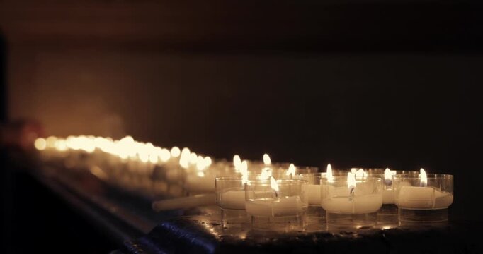 Votive candles on a marble altar in a dim church, shallow focus holds the front flames sharp while distant lights melt into blur and a soft hand places a new candle on the altar in warm candlelight