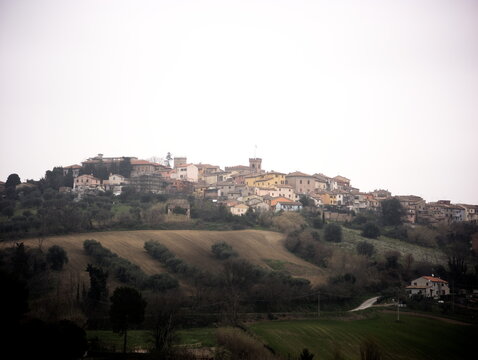 Borgo medievale italiano arroccato su una collina, circondato da campi verdi, ulivi e dolci colline. Le case in pietra con tetti in coppi rossi si raccolgono attorno alla torre storica del centro