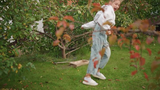 White girl dancing in backyard garden, playful twirls and small cartwheel attempt among autumn leaves casual hoodie and jeans, white sneakers on green grass, sunlight through branches, raised planter