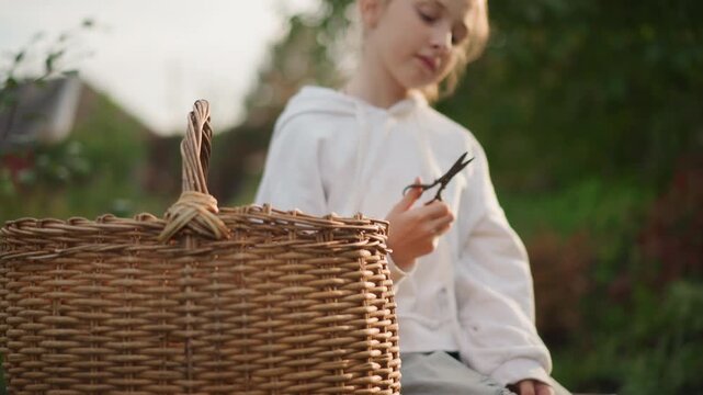 White girl outdoors with wicker basket, inspecting small scissors while sitting on garden bench in soft golden afternoon light, wearing hoodie, calm expression, focused hands preparing simple craft